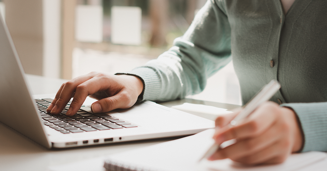 Photo of woman sitting at a computer desk and writing notes in a notebook.