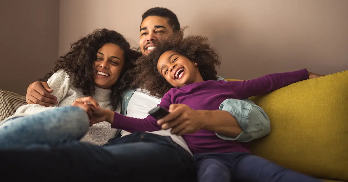 Photo of a mother and father sitting on a couch with their child and holding a TV remove, everyone is smiling. Photo by Adobe Stock.