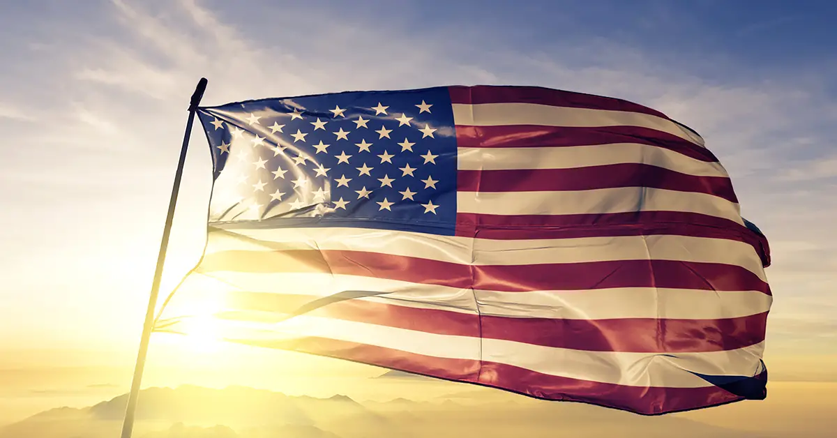 Photo of the United States flag waving in the wind, with a sunrise in the background. Photo by Adobe Stock