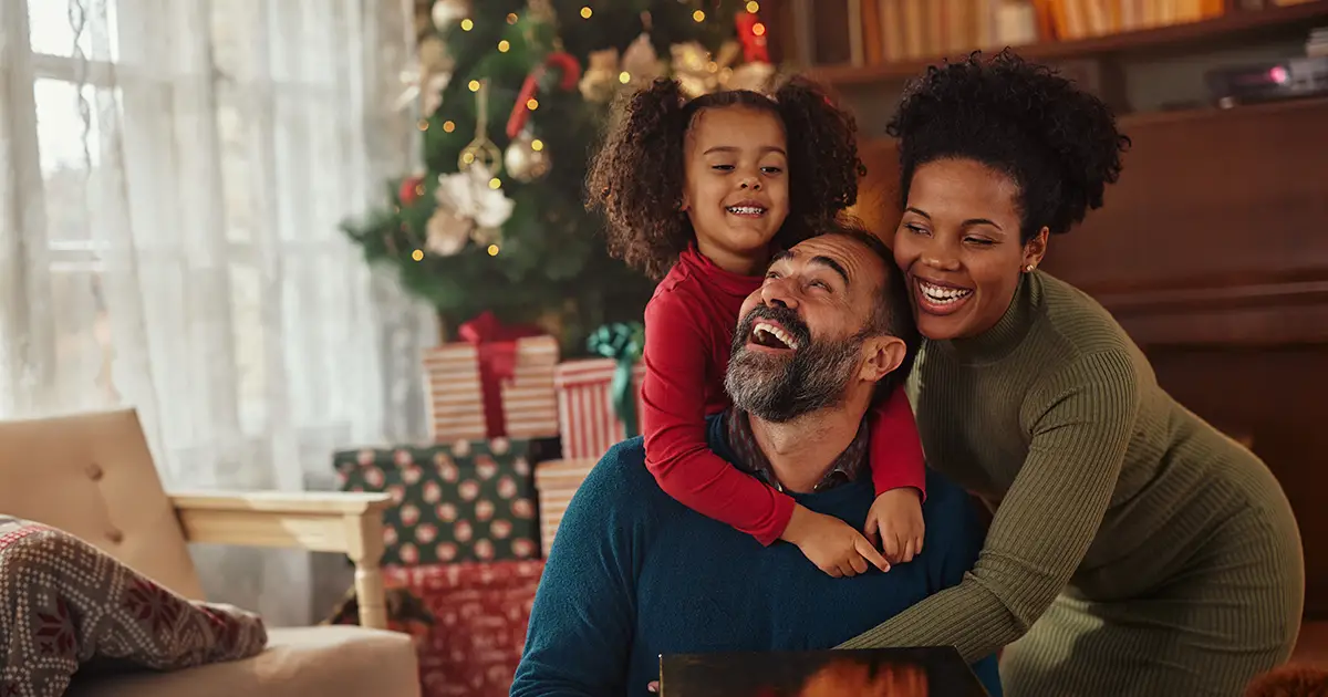 Photo of a father, mother, and daughter sitting down and smiling in front of their Christmas tree in their living room. Photo by Adobe Stock.