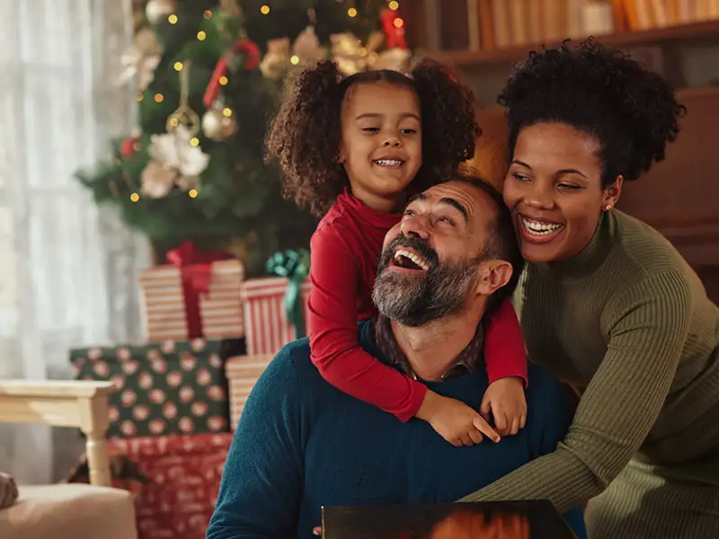 Photo of a father, mother, and daughter sitting down and smiling in front of their Christmas tree in their living room. Photo by Adobe Stock.