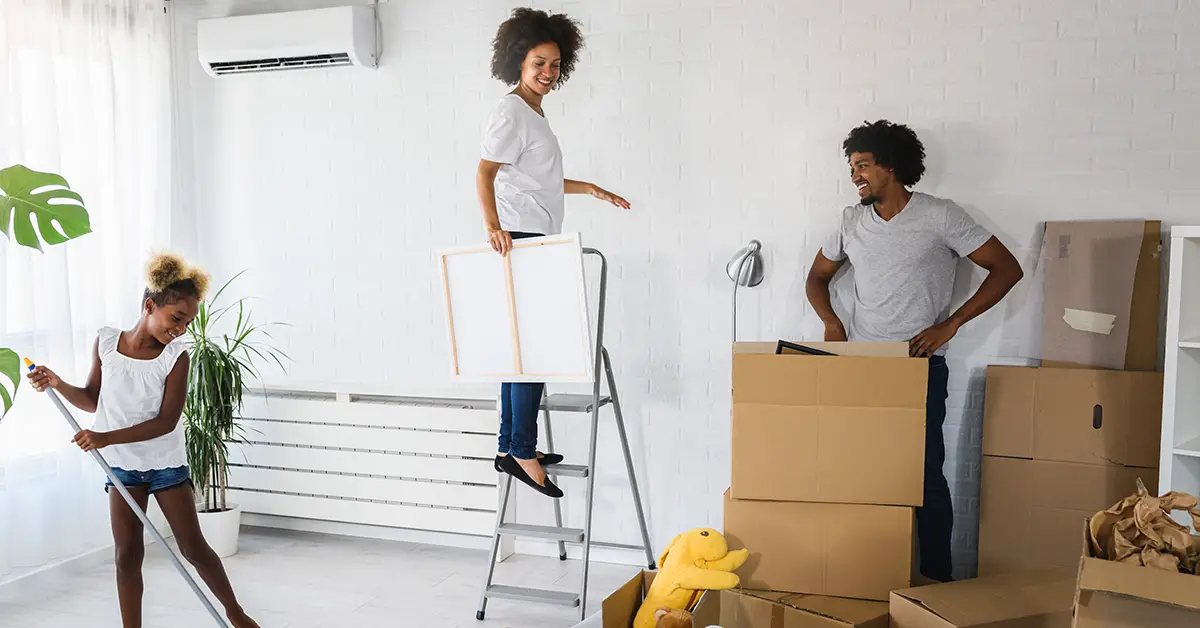 Photo of a husband, wife, and daughter in a new barren apartment doing various tasks with cardboard boxes stacked on the floor. Photo by Adobe Stock.