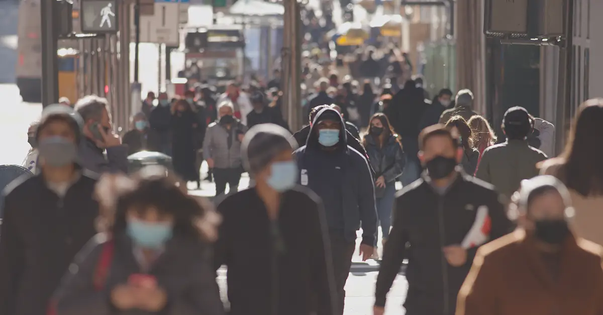 A large crowd of people wear masks on their face while walking on a city street during the COVID-19 pandemic. Photo by Adobe Stock