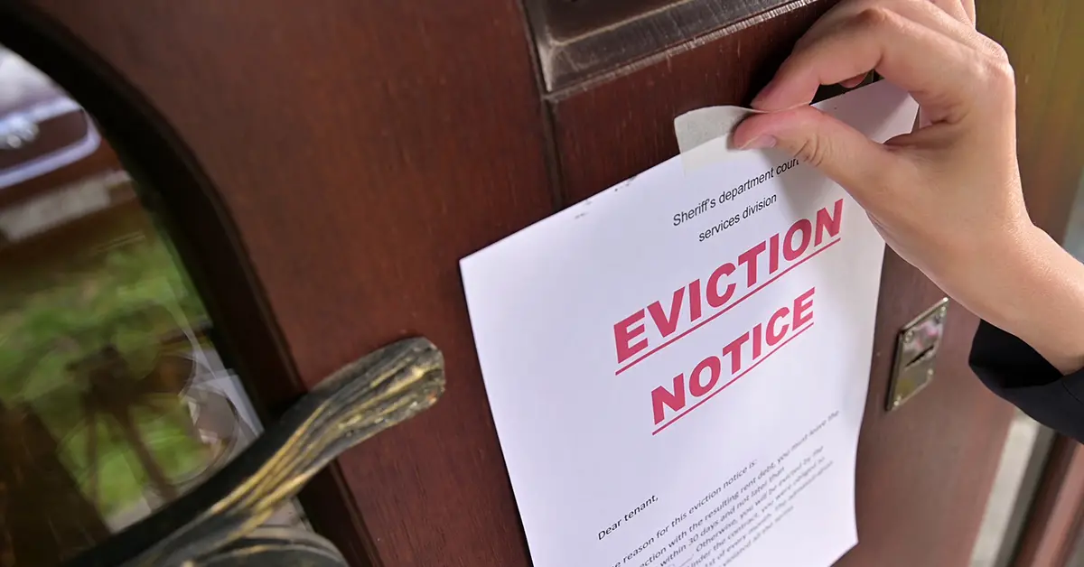 Photo of a landlord's hand taping an eviction notice letter to the front door of a rental home. Photo by Adobe Stock