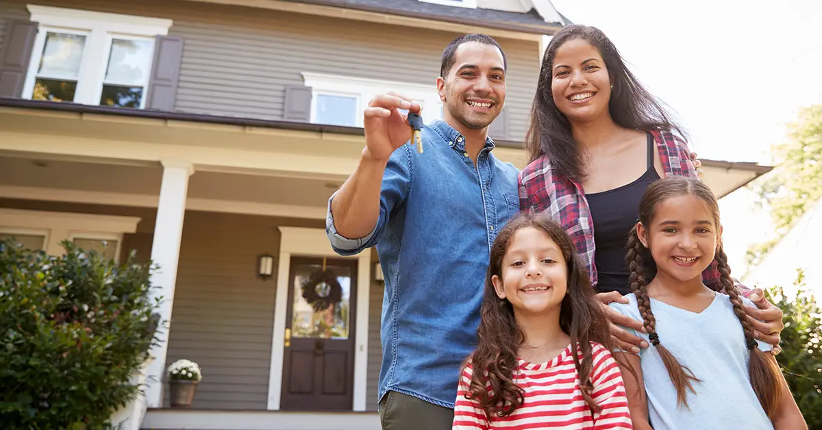 Photo of a husband and wife standing with their two daughters in front of their new rental house. As everyone smiles, the husband holds out his hand to show off the house key. Photo by Monkey Business on Adobe Stock.