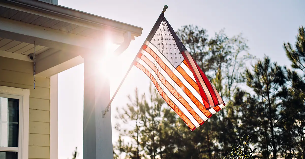 Photo of a U.S. flag mounted on a pole attached of a house, with the bright sun shining through the flag.