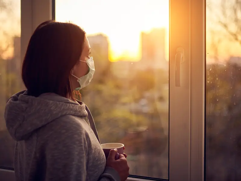 Photo of a woman with a medical mask over her face, and looking out at a sunrise through a window.