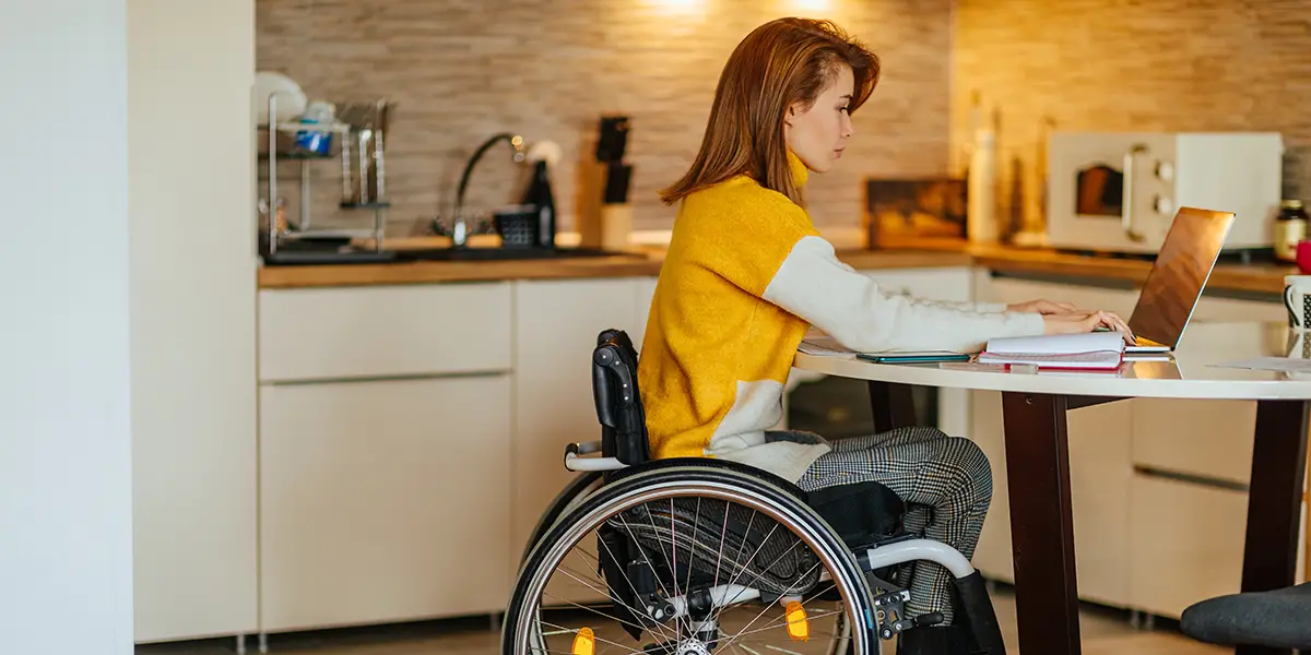 Photo of a woman in a wheelchair using her computer at her kitchen table.