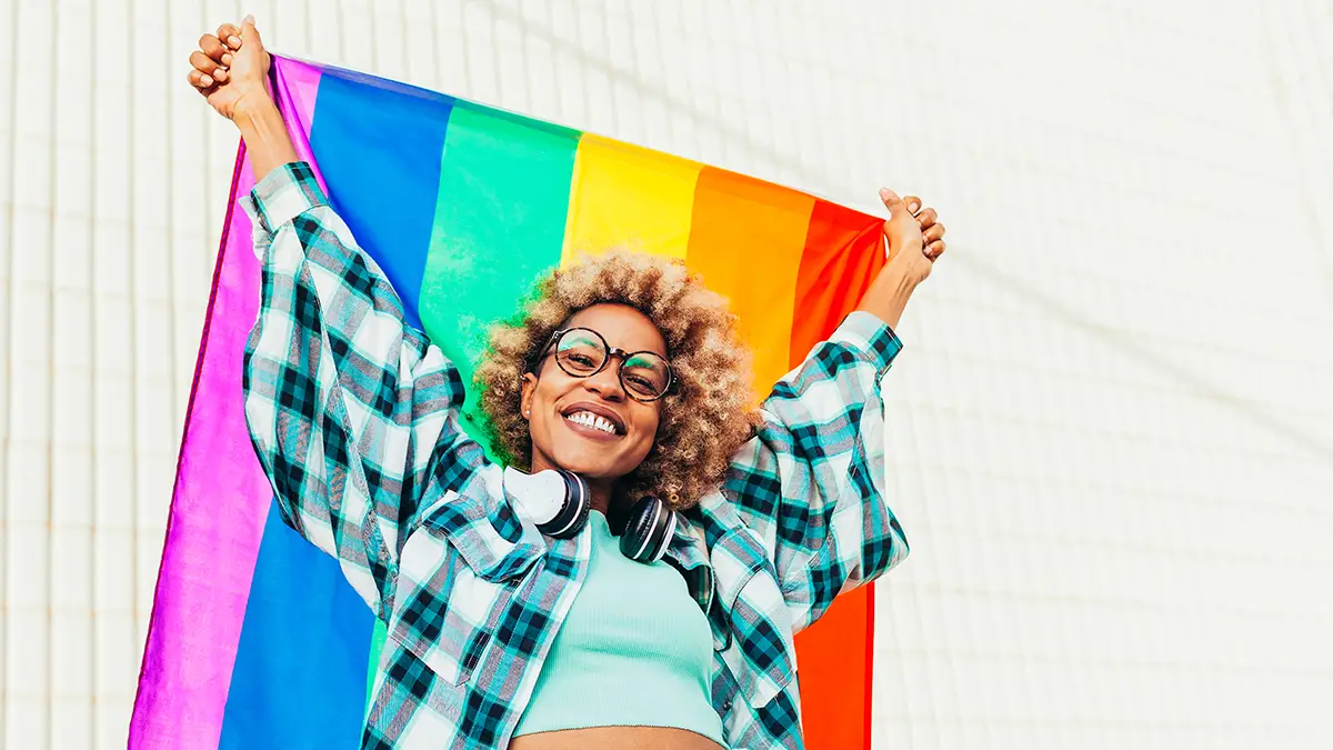 Black woman with eyeglasses, headsets and afro hair holds up the lgbtq flag of gay pride that moves with the wind on a sunny day.
