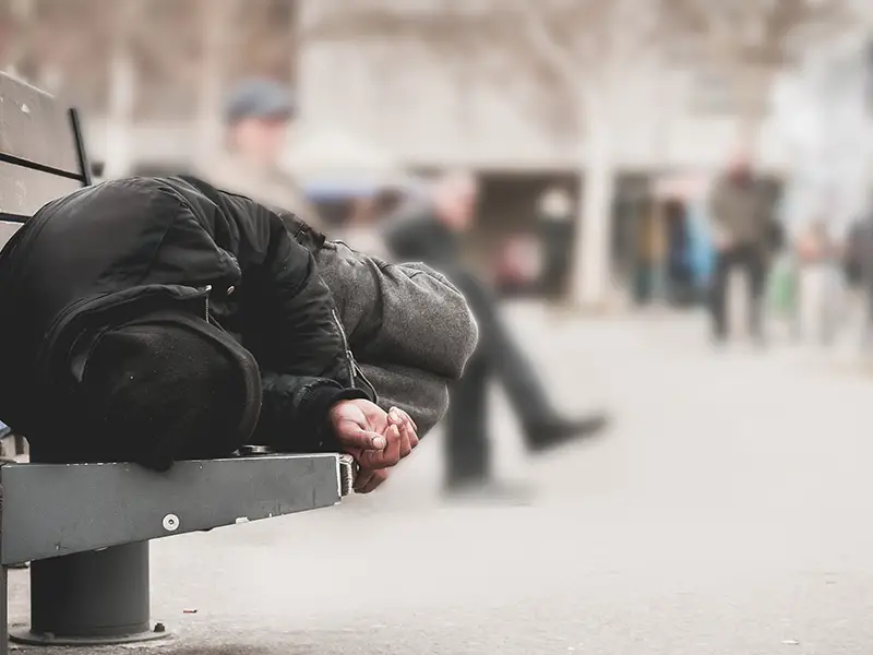Photo of a homeless person laying down on a bench during the day that's located on a busy city street.