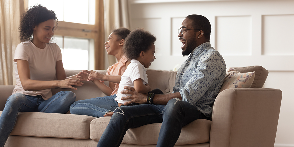 Photo of a family of four (mom, dad, and two daughters) inside their home and playing on the couch.