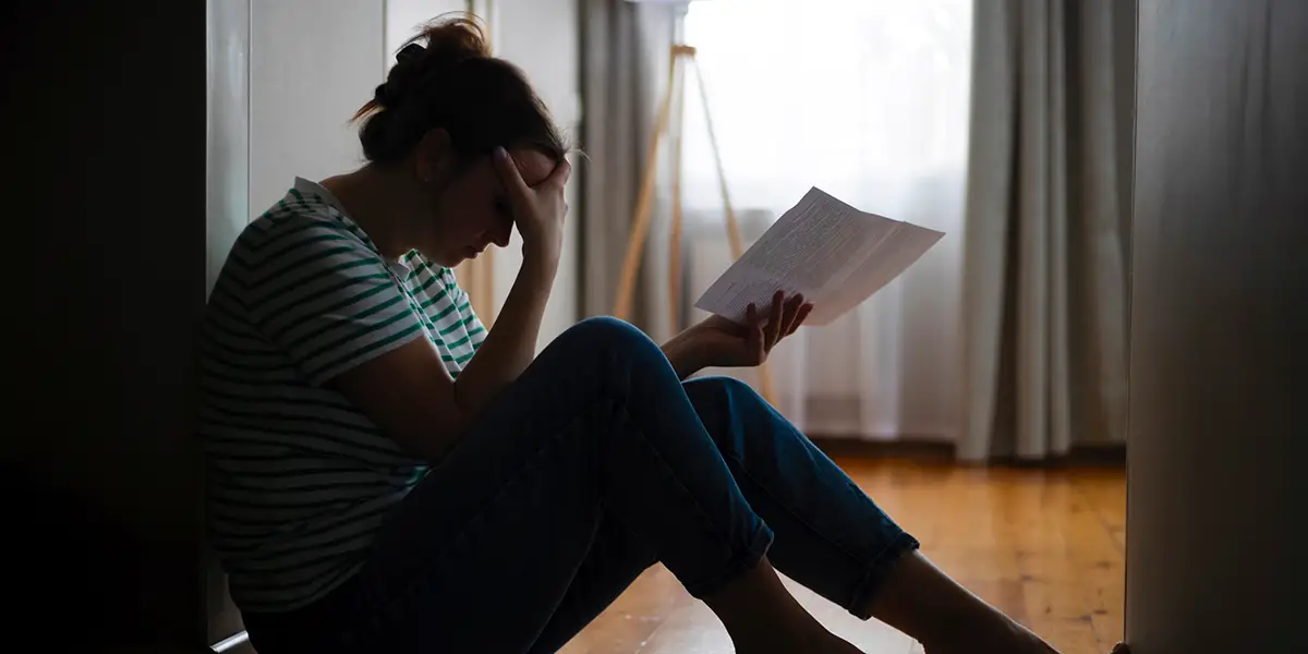 Photo of a renter sitting ont he floor of her home, with a monthly bill in one hand, and her other hand is placed on her forehead; a sign of depression.