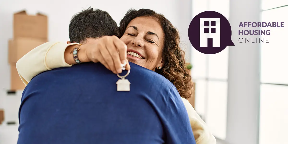 Photo of a wife and husband hugging in their new apartment. The husband's back is turned, while the wife holds the keys to their apartment in her hand, and expresses a relieving smile.