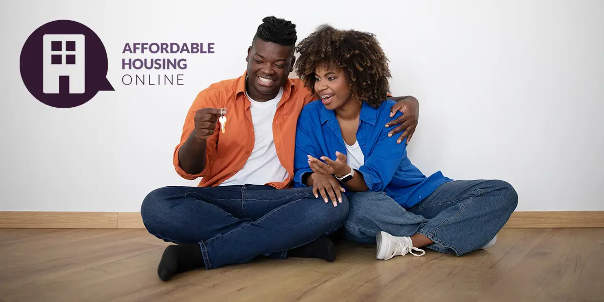 Photo of a man and woman couple sitting next to each other on the floor of their new empty apartment. Both are smiling and looking at the key to their new apartment, while the man holds up the key with one hand, and holds his significant other in the other hand. Affordable Housing Online logo appears on the top left.