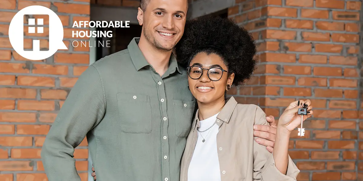 Photo of a young bi-racial couple smiling and facing the camera in front of their new apartment. The man holds the woman in his arms, as the woman holds up the keys to their home in her left hand.
