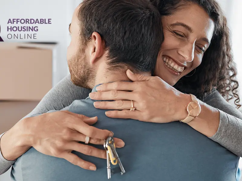 Photo of a couple hugging inside of their new apartment, with moving boxes in the background. The wife faces the camera and is smiling, as she has her arms around her husband, and keys in her right hand.