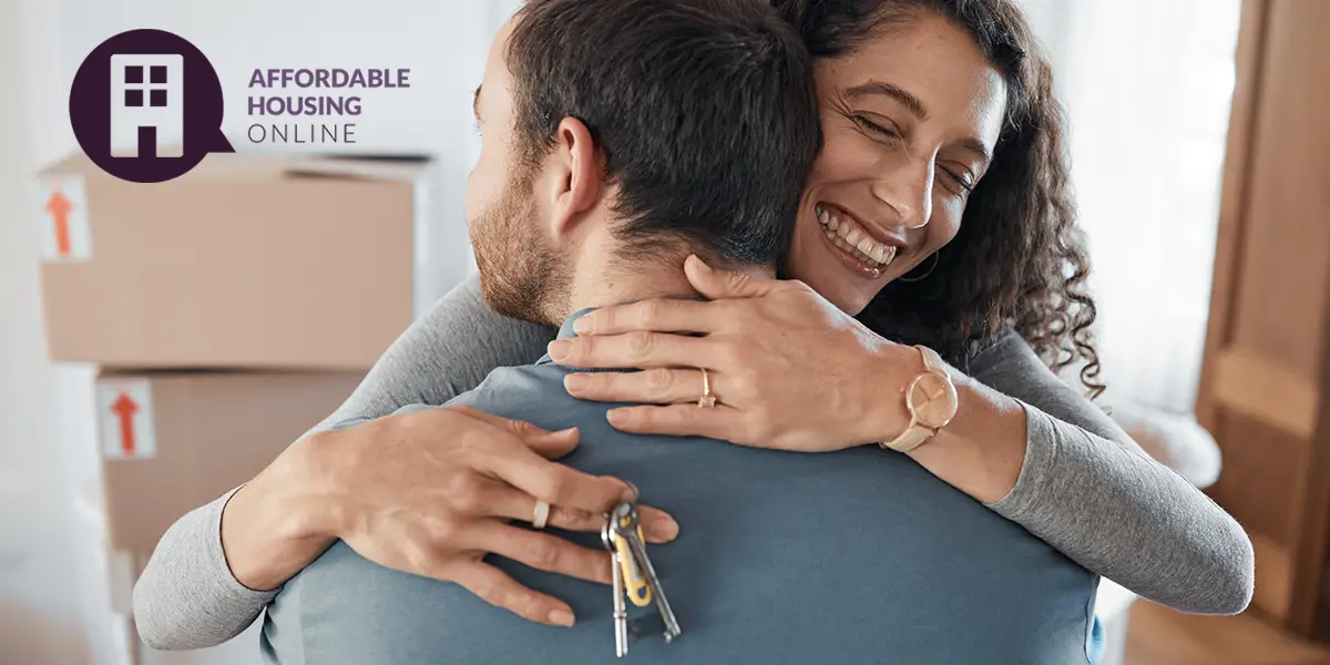 Photo of a couple hugging inside of their new apartment, with moving boxes in the background. The wife faces the camera and is smiling, as she has her arms around her husband, and keys in her right hand.