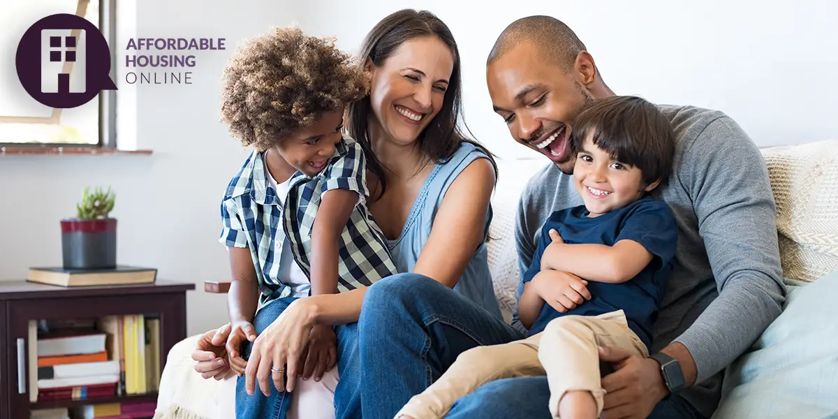 Photo of a biracial family with a mom, dad, and two sons all smiling and looking at each other. The mom and dad sit on the couch of their new home, as each son sits on their mom and dad's lap.