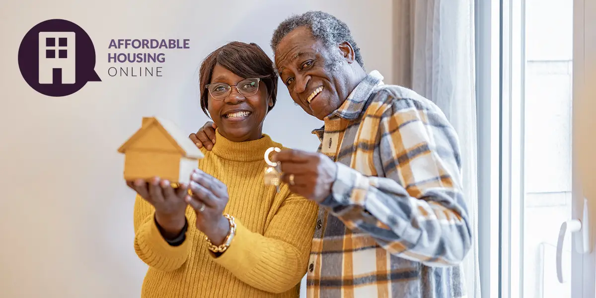 Photo of a senior couple smiling at the camera as they stand in their new home. The man on the right holds the house keys in his left hand, as his right hand embraces his wife. The woman on the left is holding out a small wooden basic home model in her hands.