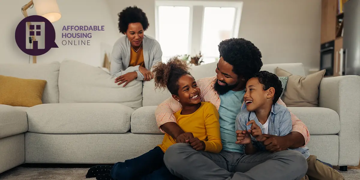 A Black family of four smile and rest as they're looking at each other in their new affordable home. The father sits on the floor, embracing his young daughter and son. And the mom smiles while leaning on the couch behind them.