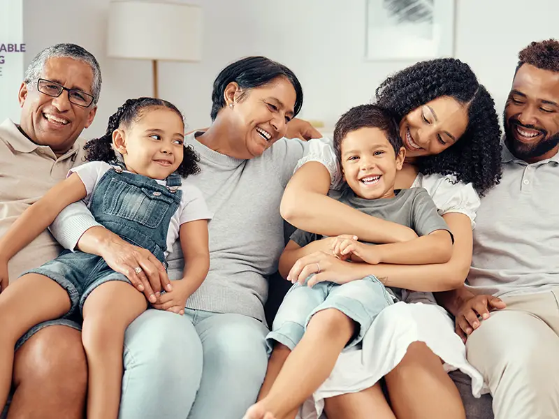 Photo of a happy multi-generational and mixed-race family sitting on the couch of their new affordable home, and smiling at each other. From left to right is the grandfather, granddaughter, grandmother, grandson, mother, and father.