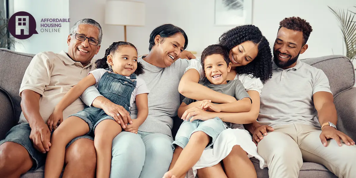 Photo of a happy multi-generational and mixed-race family sitting on the couch of their new affordable home, and smiling at each other. From left to right is the grandfather, granddaughter, grandmother, grandson, mother, and father.