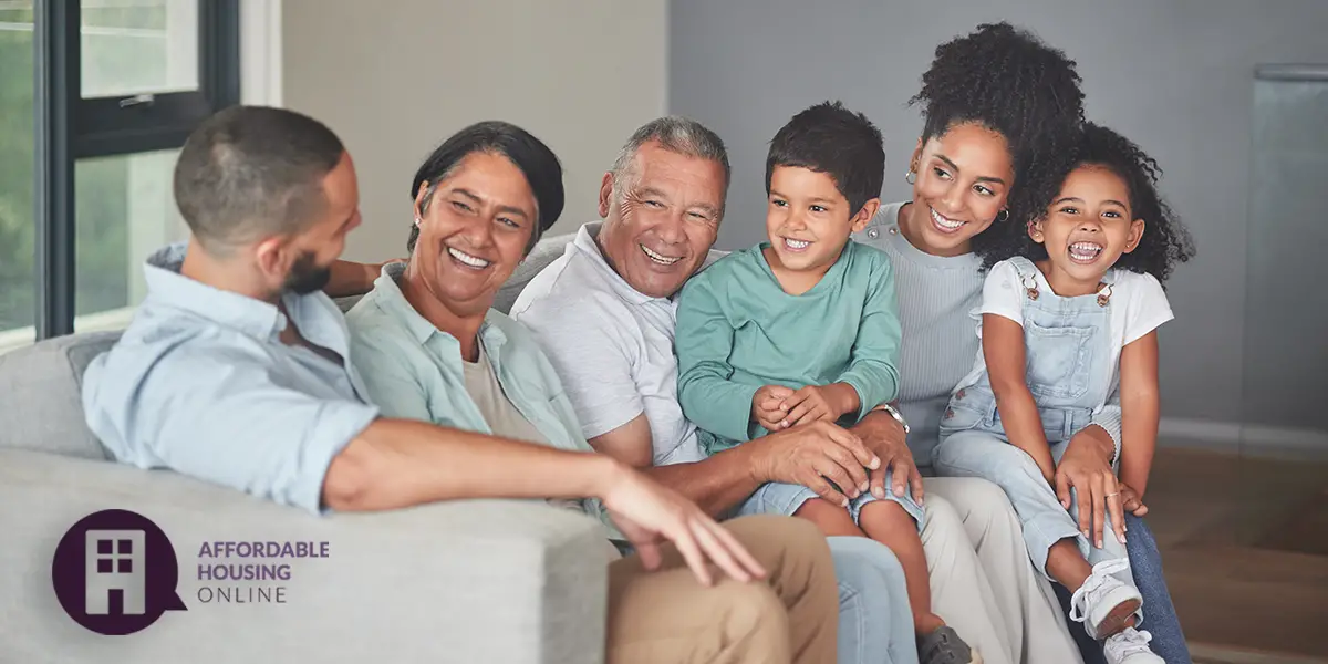 A 3-generation mixed-race family sits in the living room of their new affordable home. From left to right, a young adult man, elderly woman, elderly man, young boy, young adult woman, and young girl are all smiling and looking at each other while seated next to each other on the couch.