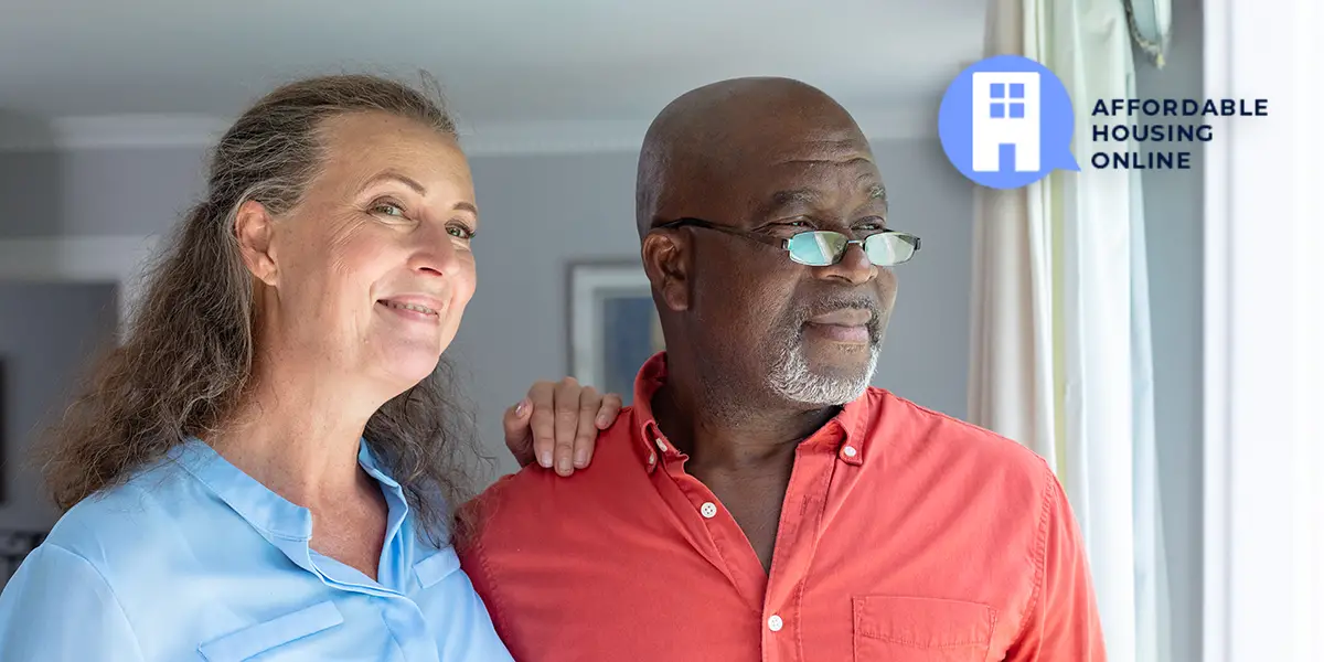 A senior bi-racial couple stands in their new affordable home. The woman on the left has her left hand on her partner's shoulder while smiling and looking at the camera. The man on the right looks out the window with a determined facial expression.