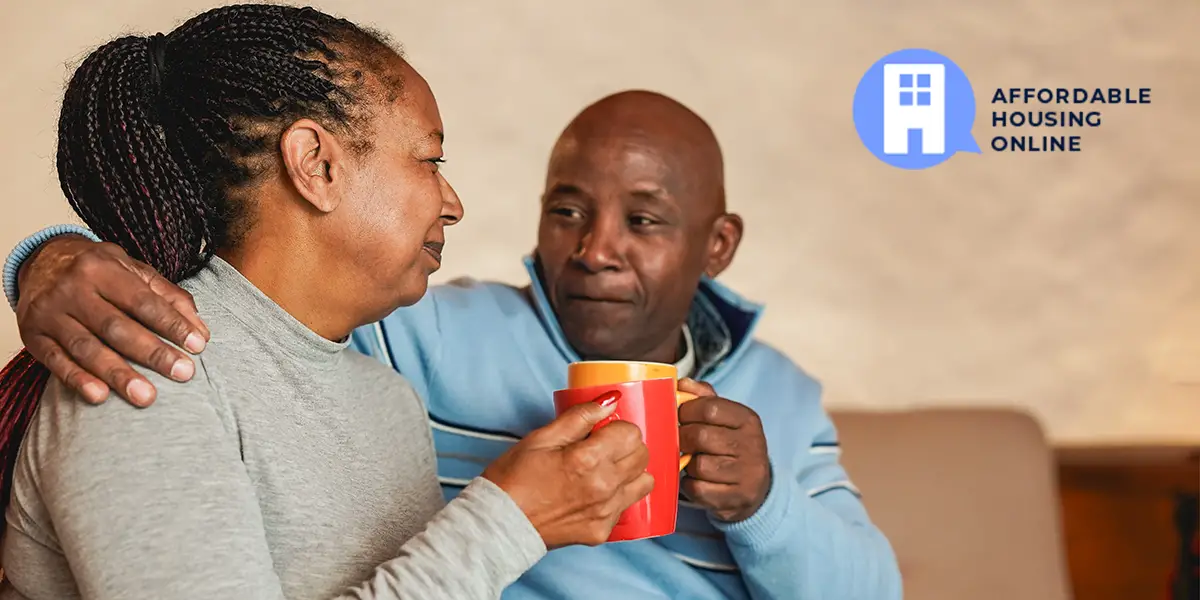 A black couple sit next to each other on the couch in their new affordable apartment, and look at each other with relieved expressions while each of them hold a coffee mug. The husband on the right holds his right arm around his wife, to the left.