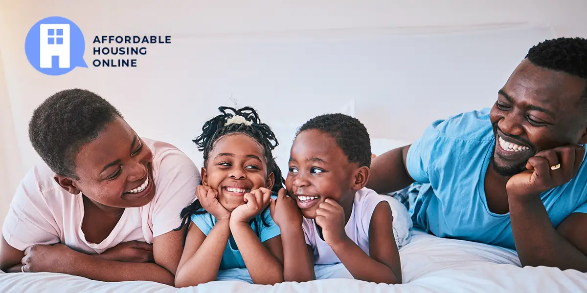 A Black family of four smiles and looks at eachother while laying on a bed in their new affordable home. From left to right, the mom looks at her daughter to the right of her, the daughter looks at her brother to the right of her, the brother looks at his sister to the left of him, and the father looks at his son to the left of him.