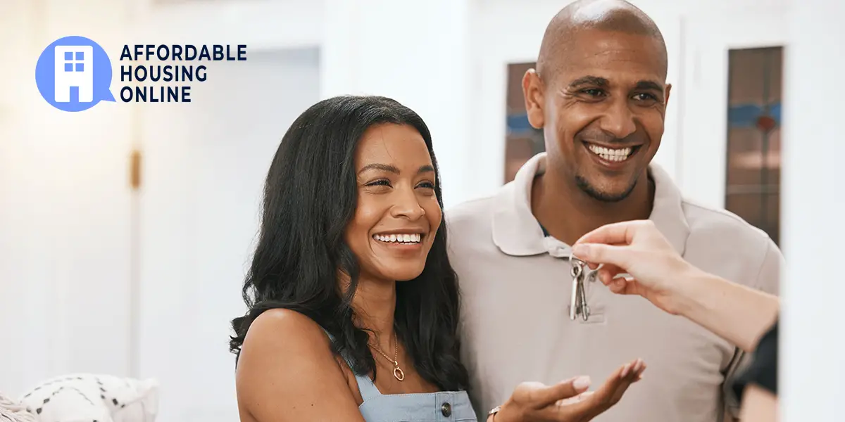 Photo of a man and woman smiling, as the woman on the left reaches out with her hand to be given keys to their new affordable home.