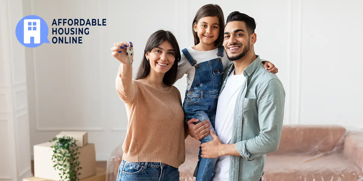 Photo of a family of a mom, dad and child smiling while standing in the living room of their new affordable home. The mom on the left holds up house keys, with the Affordable Housing Online logo to her left.