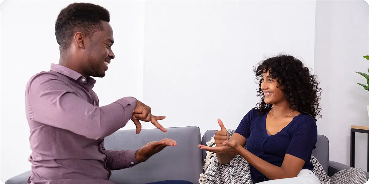 A man (left) and woman (right) face each other while communicating with sign language in a living room.