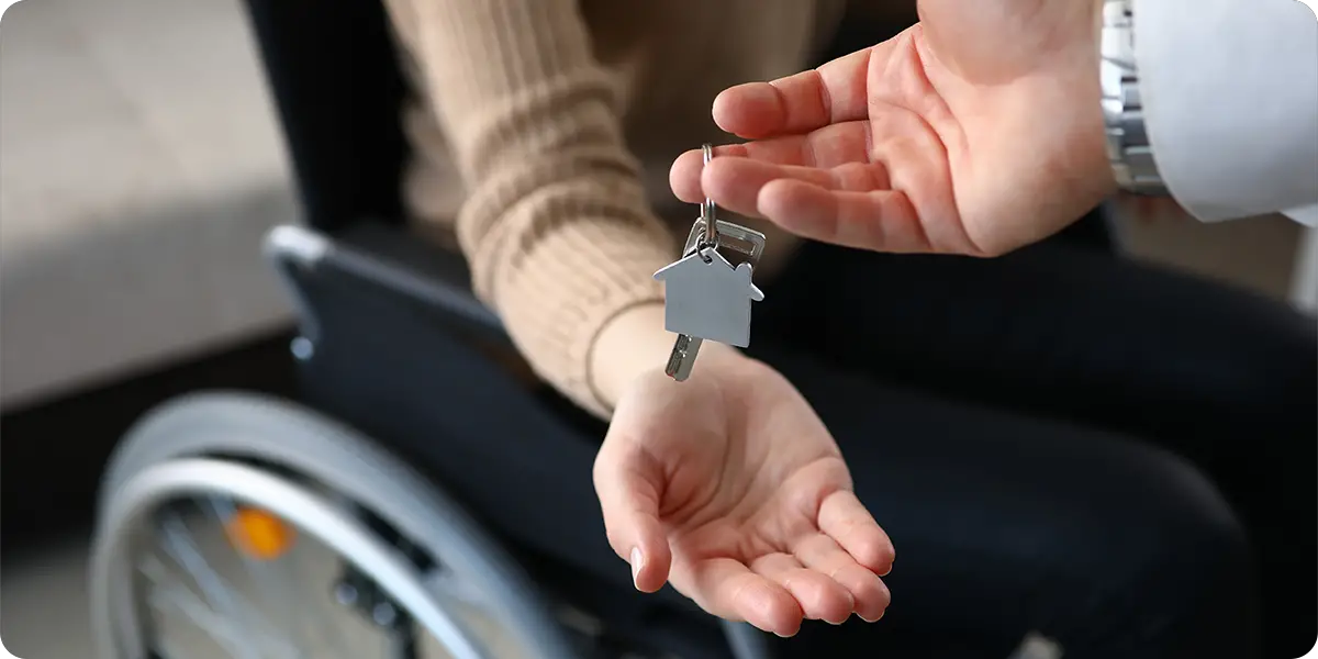 A landlord's hand on the right gives the keys to an affordable apartment to a person on the left sitting in a wheelchair, with their right hand open.