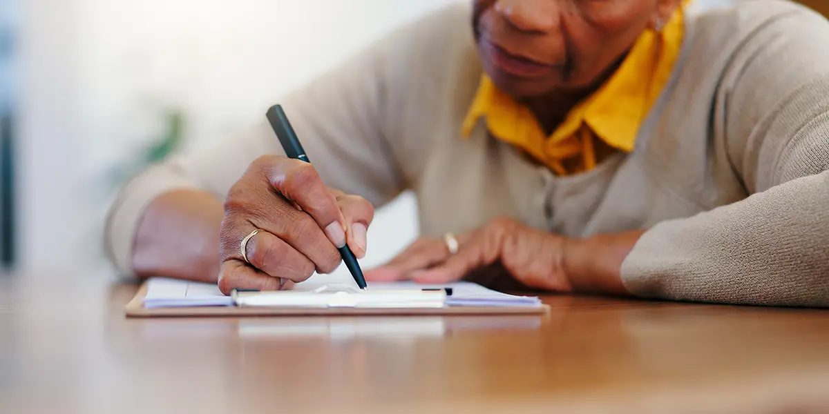 Photo of senior Black woman sitting down at a table, with a housing application in front of her on the table. A pen is in her right hand, as she fills out the application.